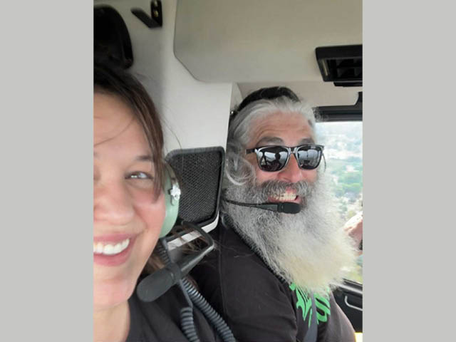 A woman with black hair smiling next to a man with a long gray beard inside a helicopter