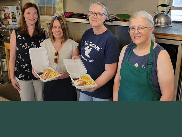 Four women standing in a kitchen, two holding takeout boxes with dinners inside