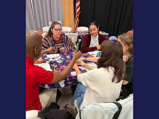 Four women and a man sitting around a table with straws and felt pipe cleaners in an upright structure