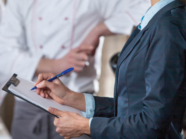 A woman with a clipboard with a person in a chef uniform in the background