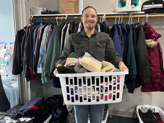 a white man holding a laundry basket of scarves in front of a full coat rack