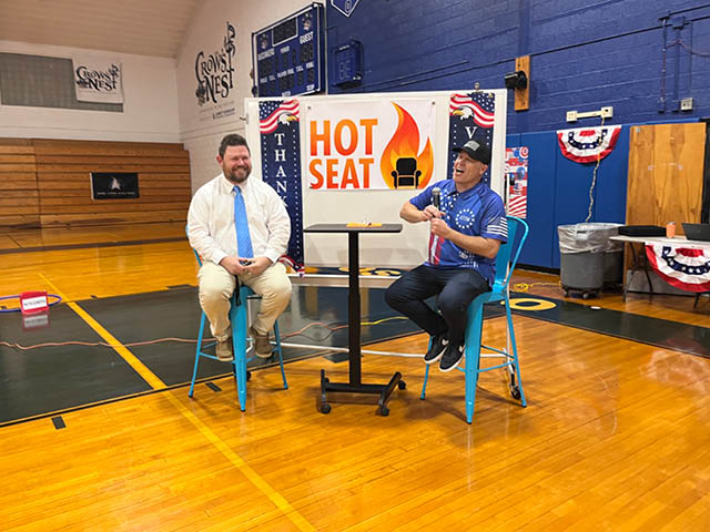 Two white men sitting in front of a display labeled "Hot Seat" in a school gymnasium