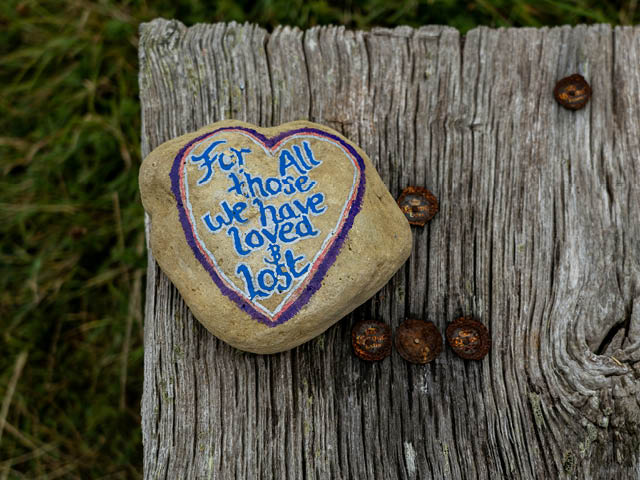 A rock on a faded wooden plank. The rock is painted with a heart and the words: For all those we have loved and lost