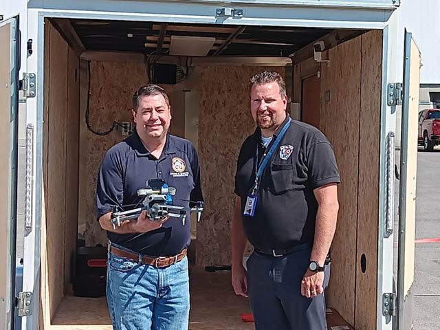 Two white men standing in front of an enclosed trailer; one of the men holds a drone
