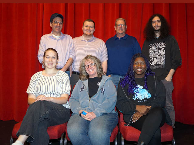 The seven cast members in front of a red curtain