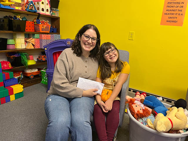 Shue and Longley sit together in a chair in the children's section of the Hannibal Free Library with an envelope containing Longley's donations. She is holding a small pumpkin and smiling.
