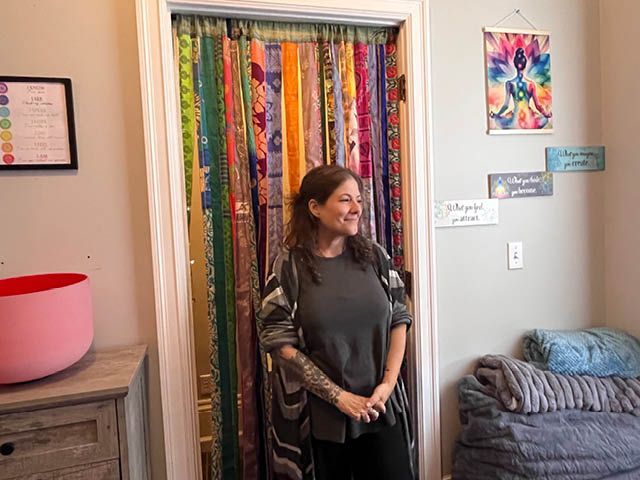 Kira Axtell in front of a curtain in Sunshine Healing; a sound bath bowl is off to the left of the image and a comfortable chair is to the right