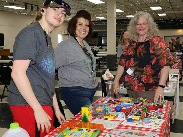 A young man and two older women stand at a cluttered craft table