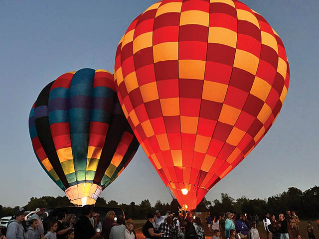 Two hot air balloons at dusk floating just over the Don Butler Fields; a crowd is visible on the ground