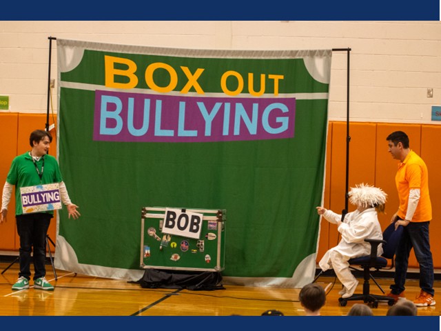 Green background with box out bullying on it and 3 people in front of it.
