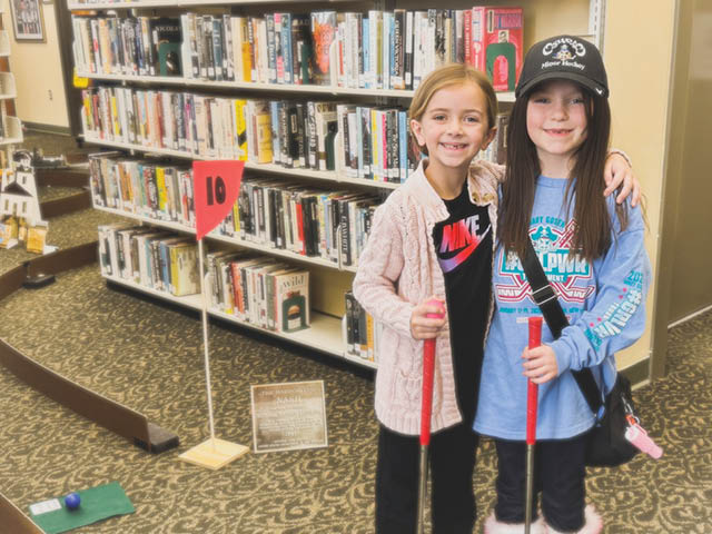 Two girls with mini golf clubs in the library smiling