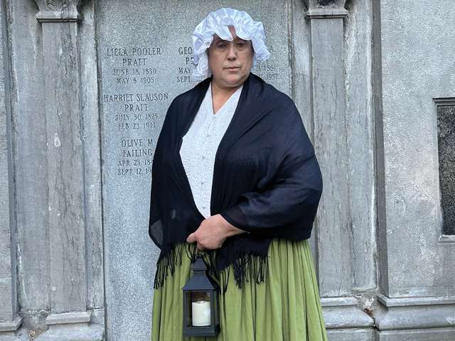 A female volunteer dressed in period clothing and holding a lantern in front of the Pratt family crypt at Mt. Adnah