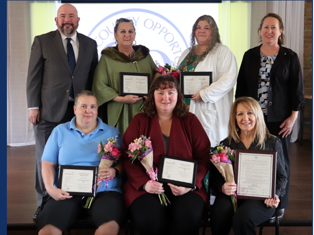Seven people, 4 standing and 3 sitting in front, middle 5 hold certificates and small bouquet of flowers