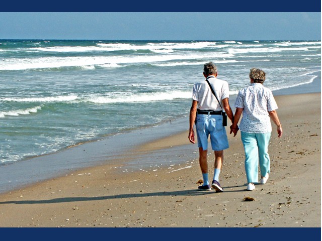 Two seniors on beach holding hands while aves wash ashore.