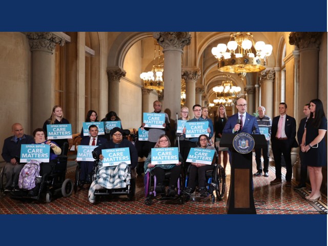 Picture shows Leader Ed Ra at the podium with about a dozen people to the left, several in wheelchairs, holding blue and white signs with 'spina bifida care matters' printed on them. On the right includes a young woman in black dress, and two others in suits,