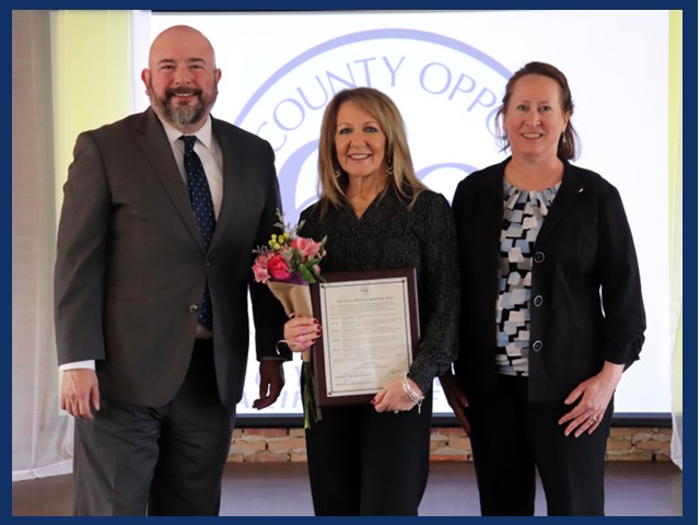 Man on left in gray suit with grayish beard, blonde woman in center in black holding small bouquet of pink flowers and an award plaque, Lady on right with dark hair tied in back, black sweater and pants with print top beneath.