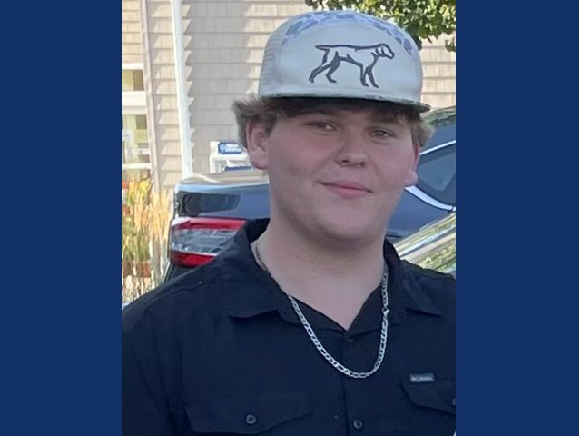 Young man with blue polo, chain on neck, with white cap with dog on it and blond waves sticking out from it.